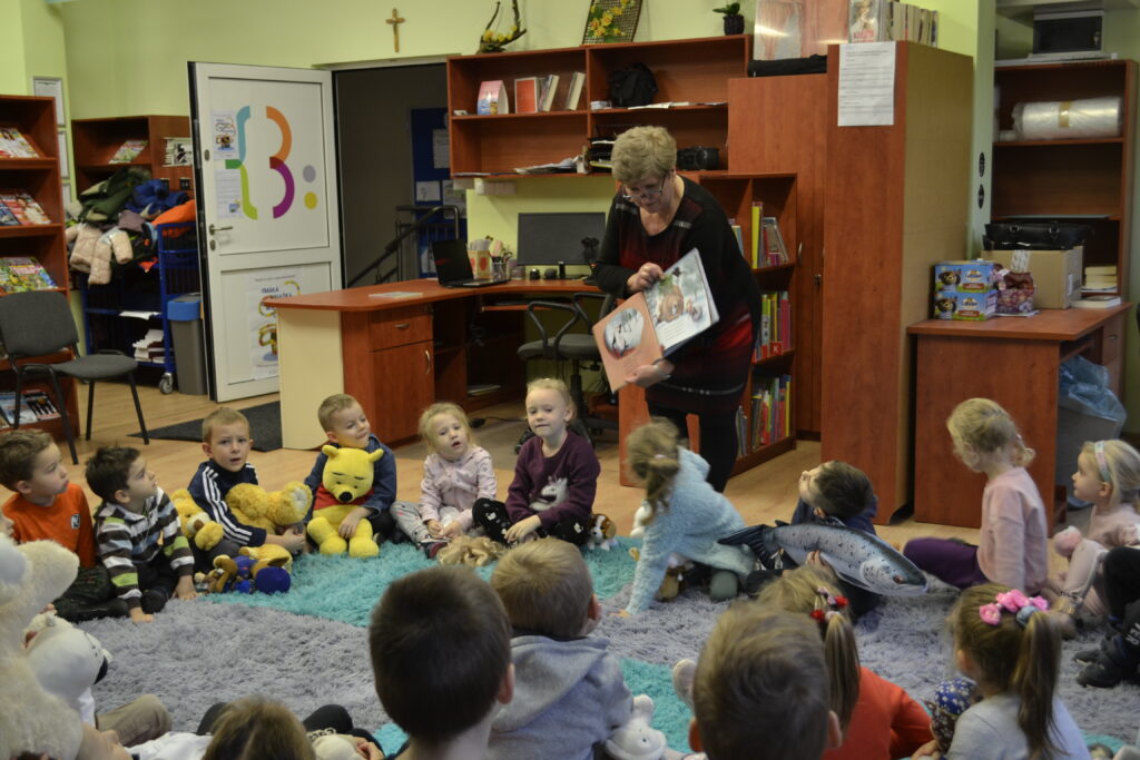 dzieci w bibliotece siedzą na dywanie i trzymają misie bibliotekarka czyta  im bajkę