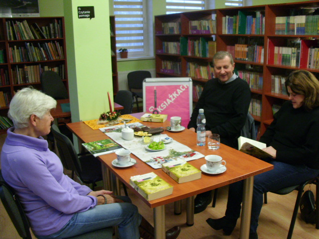 trzy osoby w bibliotece przy stole z kawą i ciastkiem  rozmawiają o książkach