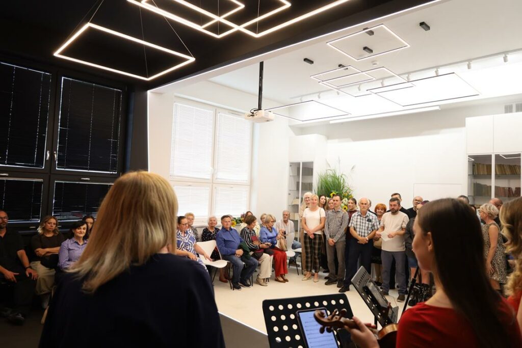 Duża grupa osób uczestniczy w wernisażu. Dyrektor biblioteki otwiera imprezę.
